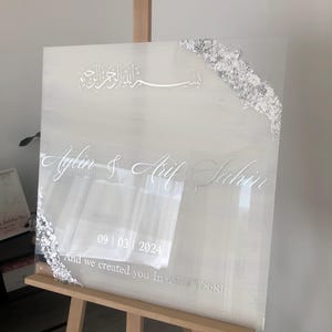 May include: A clear acrylic wedding sign with silver foil accents on the corners. The sign displays names, a date, and a quote in elegant white script. The sign is propped on a wooden easel.