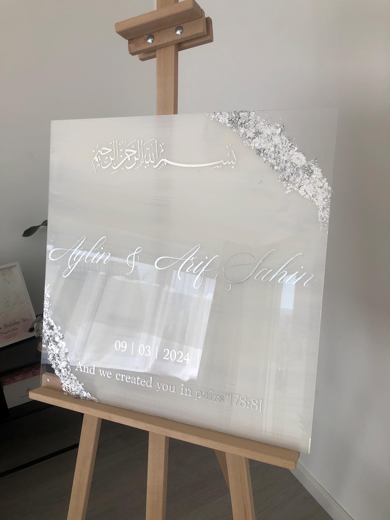 May include: A clear acrylic wedding sign with silver foil accents on the corners. The sign displays names, a date, and a quote in elegant white script. The sign is propped on a wooden easel.