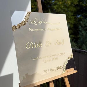May include: A beige wedding sign with gold accents, displayed on a wooden easel. The sign features gold lettering with the names "Dilek & Said", the date "30|08|2025", and a quote. Decorative gold trim adorns the edges.