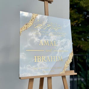 May include: A square sign with gold lettering and accents, displayed on a wooden easel. The sign features Arabic script and English text that reads "AMAL and IBRAHIM" with a quote from the Quran. The background has a cloudy sky design.