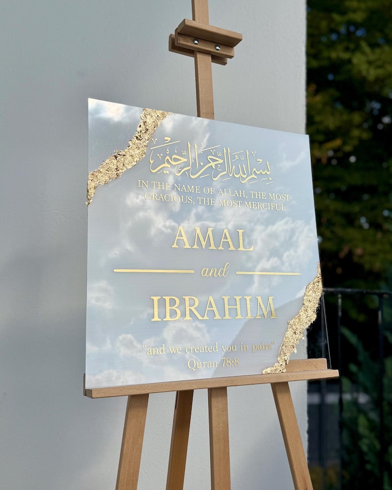 May include: A square sign with gold lettering and accents, displayed on a wooden easel. The sign features Arabic script and English text that reads "AMAL and IBRAHIM" with a quote from the Quran. The background has a cloudy sky design.