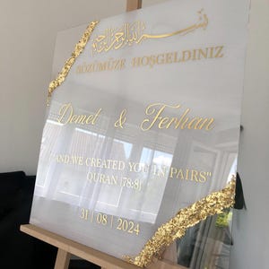 May include: A white acrylic wedding sign with gold lettering and accents. The sign includes the names Demet & Ferhan, the date 31/08/2024, and a quote from the Quran. Gold leaf decorates the edges.
