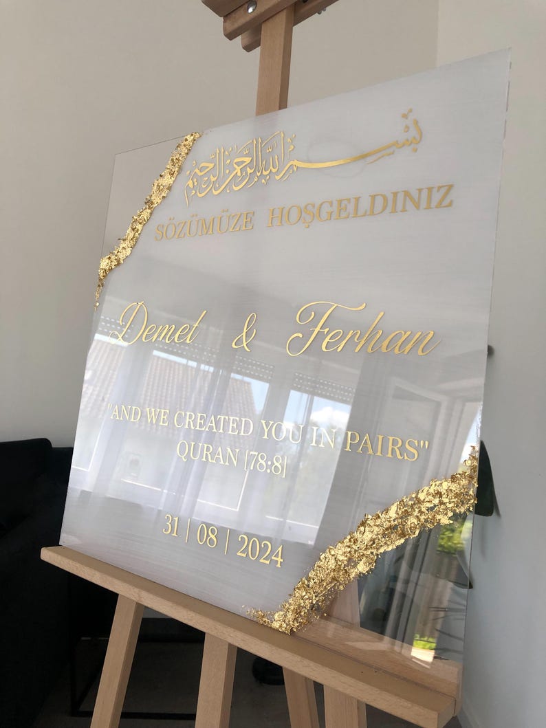 May include: A white acrylic wedding sign with gold lettering and accents. The sign includes the names Demet & Ferhan, the date 31/08/2024, and a quote from the Quran. Gold leaf decorates the edges.