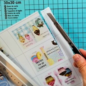 May include: A close-up of a sheet of paper with printed images of ice cream, donuts, and cupcakes. The paper is being cut with a utility knife on a blue cutting mat. The text on the paper includes "The book of ice creams", "Donuts of all colors", and "How to make homemade fluffy cupcakes".