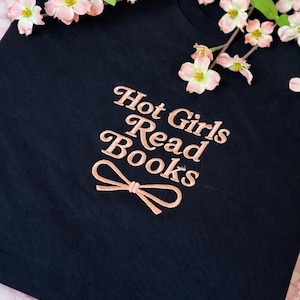 Könnte beinhalten: Ein marineblaues T-Shirt mit dem gestickten Schriftzug "Hot Girls Read Books" in einem hellen Pfirsichton, zusammen mit einem Schleifendesign. Das Shirt ist mit zarten rosa und weißen Blüten geschmückt.