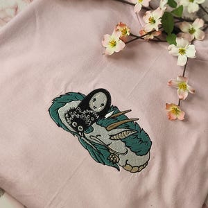 Könnte beinhalten: Ein hellrosa T-Shirt mit einem gestickten Design einer Figur aus einem japanischen Zeichentrickfilm. Das Design umfasst eine schwarze Figur mit weißem Gesicht und ein grün-weißes Wesen. Rosa und weiße Blumen sind im Hintergrund.