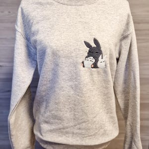 Könnte beinhalten: Ein graues Sweatshirt mit einem gestickten Bild eines schwarz-weißen Totoro-Charakters, der einen kleineren weißen Totoro-Charakter hält.