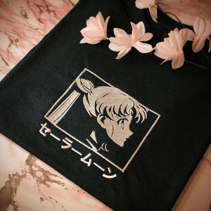 Könnte beinhalten: Dunkelblaues T-Shirt mit silberner Stickerei von Sailor Moon und japanischem Text. Das T-Shirt ist mit rosa Blüten verziert.
