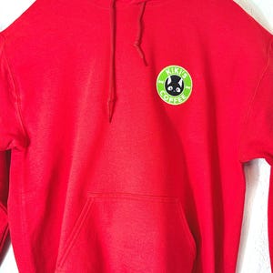 Könnte beinhalten: Ein leuchtend roter Kapuzenpullover mit einer Kängurutasche auf der Vorderseite und passenden roten Kordelzügen. Auf der linken Brust befindet sich ein runder Aufnäher mit lindgrünem Rand, einer schwarzen Katzenillustration und dem Text KIKI'S COFFEE.