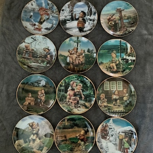 May include: A set of 12 decorative plates featuring a variety of scenes with children in traditional clothing. The plates are round and have a gold rim. Each plate depicts a different scene, such as children playing in a snowy forest, children playing in a field, and children playing in a village.