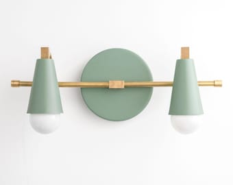 May include: A modern wall sconce with a sage green and brass finish. The sconce features two cone-shaped light fixtures with white bulbs, connected by a brass rod to a circular backplate. The design is minimalist and stylish.