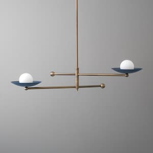 May include: A brass chandelier featuring a central vertical rod with two horizontal arms extending in opposite directions. Each arm holds a dark blue shallow bowl with a white spherical light bulb resting inside, creating a balanced mobile design.