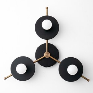 May include: A modern wall sconce with three black circular shades and brass arms. Each shade has a white globe light bulb in the center.