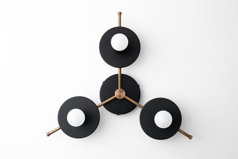 May include: A modern wall sconce with three black circular shades and brass arms. Each shade has a white globe light bulb in the center.