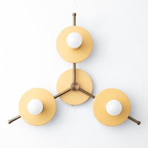 May include: A three-light wall sconce with a modern geometric design. The sconce has three circular yellow shades with white light bulbs and brass arms.