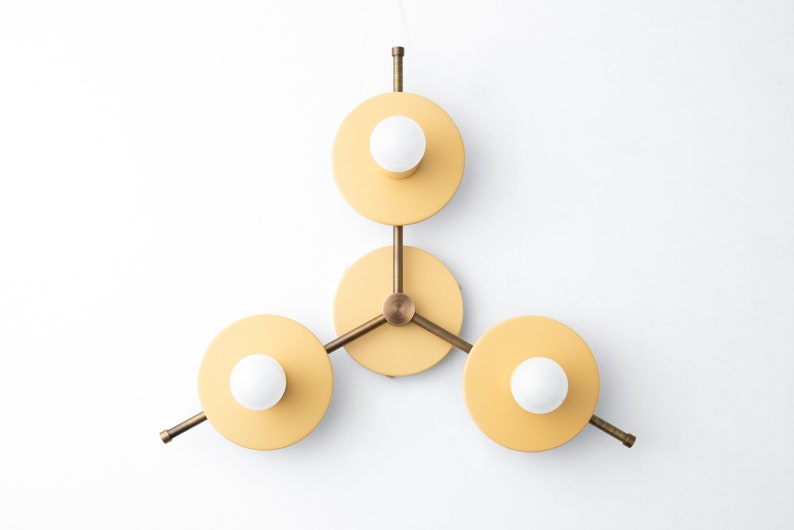 May include: A three-light wall sconce with a modern geometric design. The sconce has three circular yellow shades with white light bulbs and brass arms.
