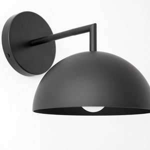 Puede incluir: Una l&aacute;mpara de pared negra con una pantalla en forma de c&uacute;pula. La pantalla es de metal y tiene un acabado liso y mate.
