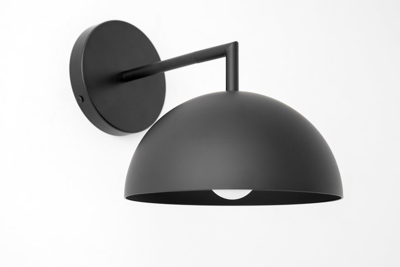 Puede incluir: Una l&aacute;mpara de pared negra con una pantalla en forma de c&uacute;pula. La pantalla es de metal y tiene un acabado liso y mate.
