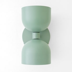 May include: A mint green wall sconce with a unique hourglass shape. The light fixture has a rounded top and bottom, connected by a circular plate and a short cylindrical piece. The sconce is mounted on a white wall.