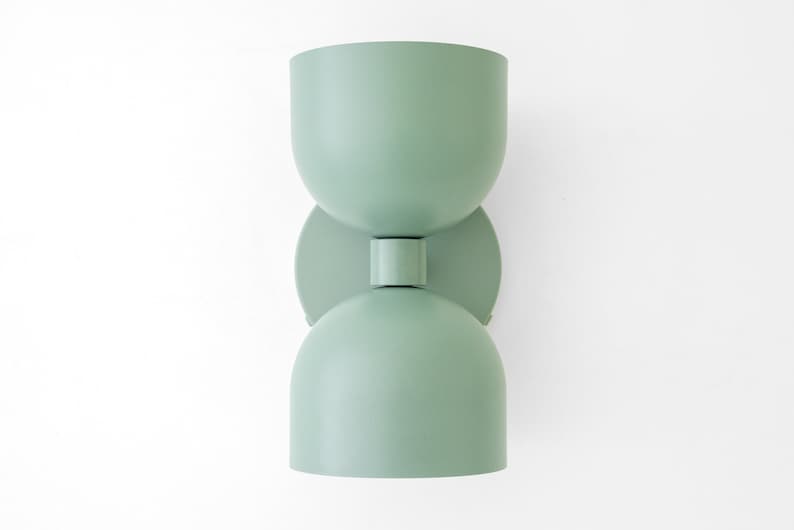 May include: A mint green wall sconce with a unique hourglass shape. The light fixture has a rounded top and bottom, connected by a circular plate and a short cylindrical piece. The sconce is mounted on a white wall.