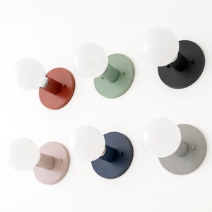 May include: A collection of six modern wall sconces. Each sconce features a round, colored base and a cylindrical arm supporting a round, white light bulb. Colors include red, green, black, pink, navy blue, and gray. The sconces are mounted on a white wall.