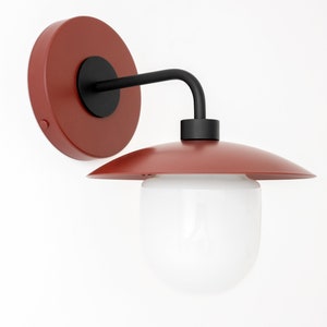 May include: A wall sconce featuring a circular terracotta-colored wall mount, a black metal arm, a matching terracotta-colored shade, and a rounded white glass globe diffuser. The fixture combines matte red-toned metal components with a smooth white glass element to create a functional lighting piece for indoor wall installation.