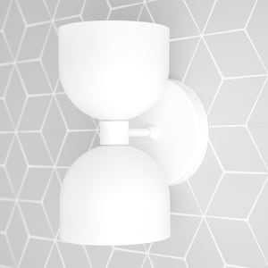 May include: A modern, white wall sconce with a double-dome design. The light fixture is mounted on a round base and features a geometric pattern on the wall behind it. The sconce is made of a smooth, matte material.