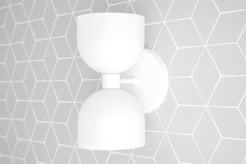 May include: A modern, white wall sconce with a double-dome design. The light fixture is mounted on a round base and features a geometric pattern on the wall behind it. The sconce is made of a smooth, matte material.