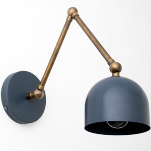 May include: A wall-mounted lamp with a navy blue dome-shaped shade and a brass-colored adjustable arm. The lamp has a circular base and a visible lightbulb. The design is modern and minimalist.