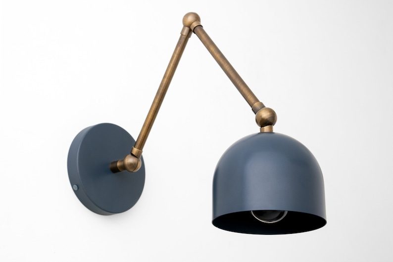 May include: A wall-mounted lamp with a navy blue dome-shaped shade and a brass-colored adjustable arm. The lamp has a circular base and a visible lightbulb. The design is modern and minimalist.