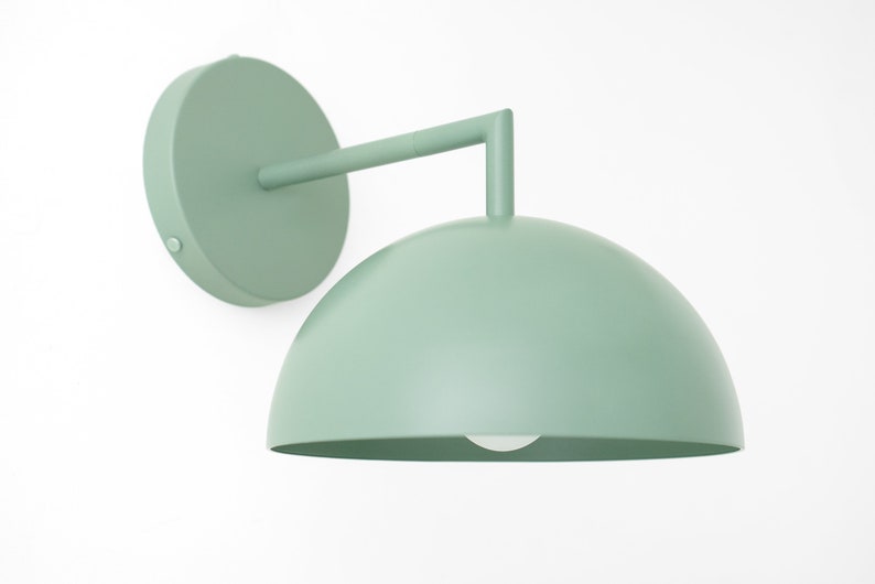 Puede incluir: Una l&aacute;mpara de pared de color verde salvia con una pantalla en forma de c&uacute;pula y una placa de pared redonda. La l&aacute;mpara tiene un dise&ntilde;o simple y moderno.