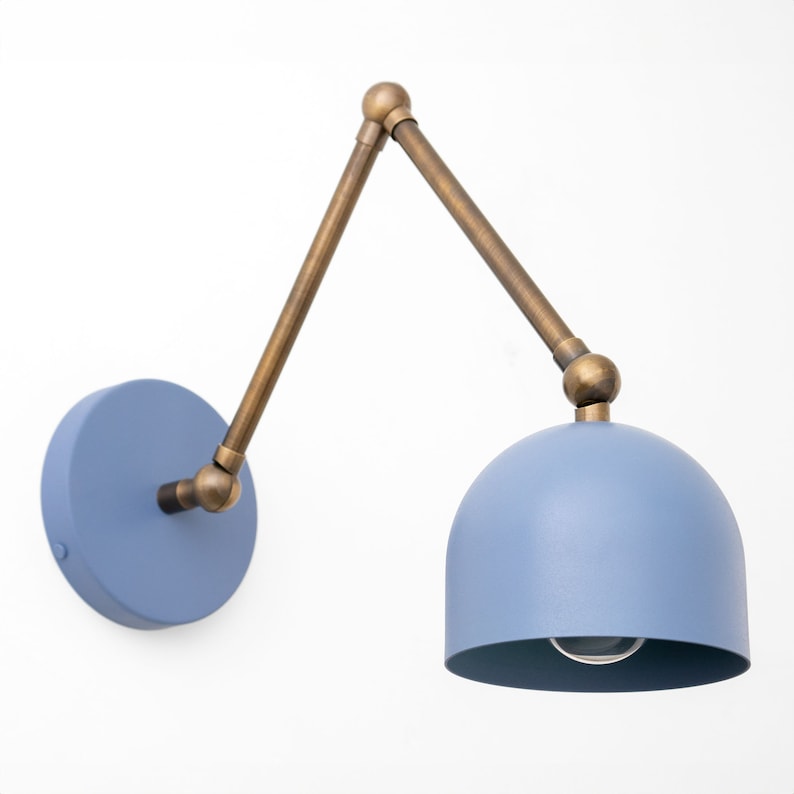 May include: A blue wall-mounted lamp with a brass-colored adjustable arm and a rounded lampshade. The lamp has a circular base and a single light bulb visible inside the shade. The design is modern and minimalist.