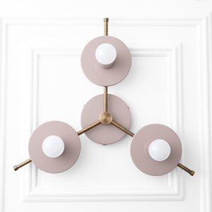 May include: A three-light wall sconce with a modern design. The sconce has a brass frame and three circular light fixtures with a dusty rose finish. Each light fixture has a white globe light bulb.