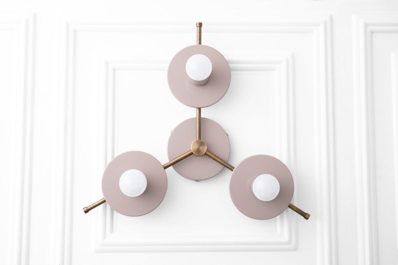 May include: A three-light wall sconce with a modern design. The sconce has a brass frame and three circular light fixtures with a dusty rose finish. Each light fixture has a white globe light bulb.