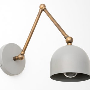 May include: A gray wall-mounted lamp with a brass-colored adjustable arm and a dome-shaped shade. The lamp has a circular base and a visible lightbulb. The arm extends from the base to the shade, allowing for directional lighting.