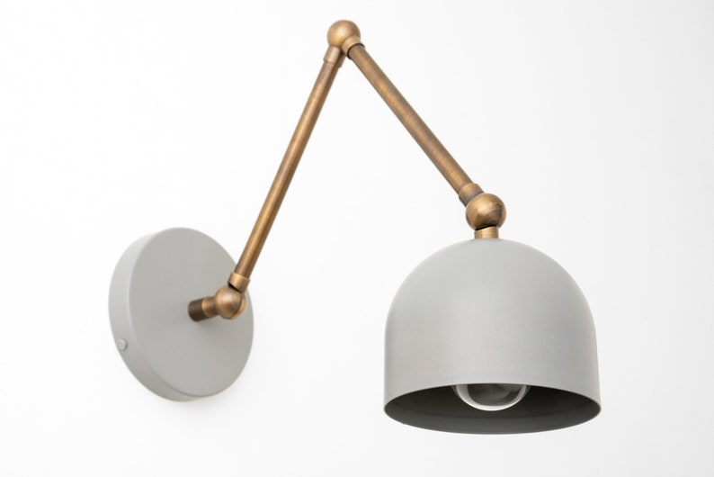 May include: A gray wall-mounted lamp with a brass-colored adjustable arm and a dome-shaped shade. The lamp has a circular base and a visible lightbulb. The arm extends from the base to the shade, allowing for directional lighting.