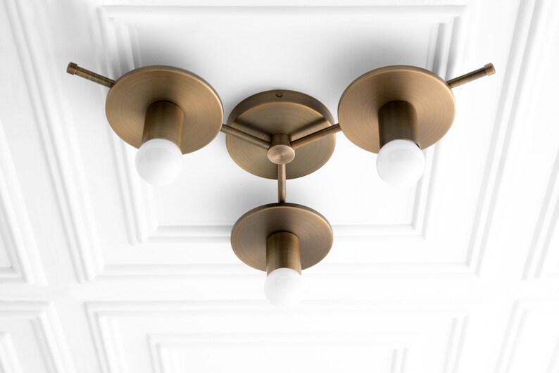 May include: A brass ceiling light fixture with three arms and three round shades. Each arm has a light bulb and a small, round, decorative piece at the end.