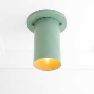 May include: A light green ceiling fixture with a cylindrical shade and a round base. The shade has a gold interior and emits a warm glow.