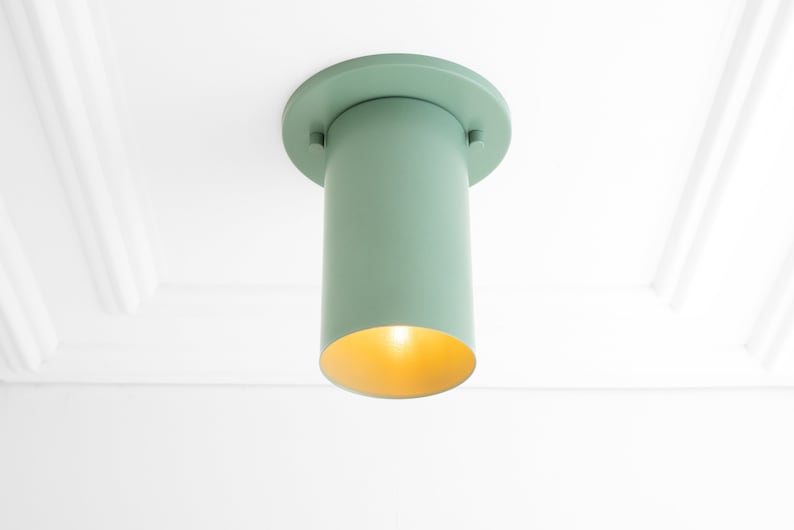 May include: A light green ceiling fixture with a cylindrical shade and a round base. The shade has a gold interior and emits a warm glow.