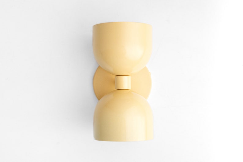 May include: A light yellow wall sconce with a unique hourglass shape. The sconce has two rounded, bowl-like shades connected by a short, cylindrical stem. It is mounted on a circular backplate.