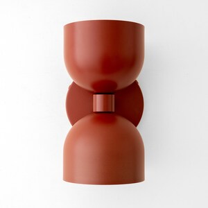 May include: A modern, terracotta-colored wall sconce. The light fixture has a unique hourglass shape, with two rounded cup-like shades connected by a circular disc and a cylindrical base. The sconce is mounted on a white wall.