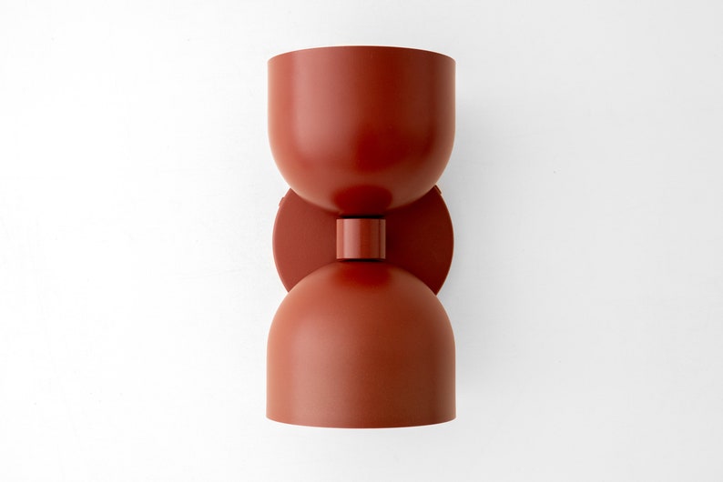 May include: A modern, terracotta-colored wall sconce. The light fixture has a unique hourglass shape, with two rounded cup-like shades connected by a circular disc and a cylindrical base. The sconce is mounted on a white wall.