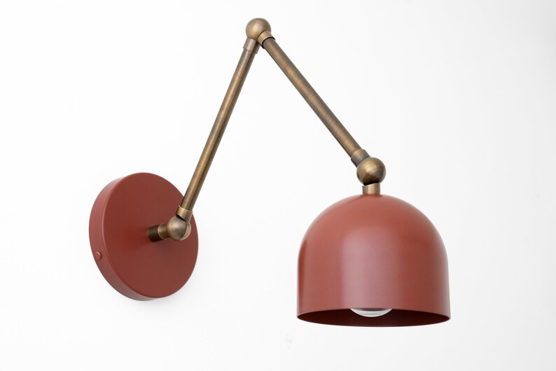 May include: A wall-mounted, adjustable lamp with a rust-colored dome shade and a circular base. The lamp features a brass-toned, articulated arm and a small bulb. The design is modern and suitable for various interior styles.