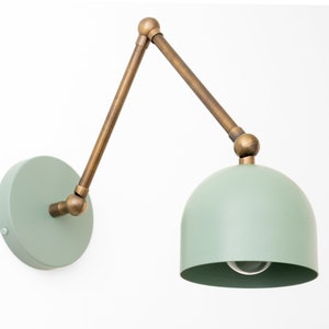 May include: A wall-mounted lamp with a mint green dome-shaped shade and a circular base. The adjustable arm and connecting joints are a brass color. The lamp is designed for indoor use and is mounted on a white wall.