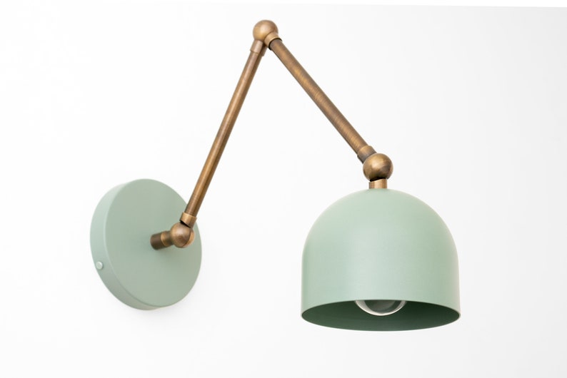 May include: A wall-mounted lamp with a mint green dome-shaped shade and a circular base. The adjustable arm and connecting joints are a brass color. The lamp is designed for indoor use and is mounted on a white wall.