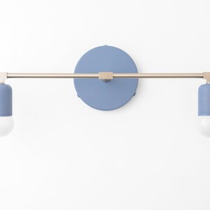 Puede incluir: Una l&aacute;mpara de pared con dos bombillas. La l&aacute;mpara tiene un brazo de metal plateado y una base redonda de color azul claro. Las bombillas est&aacute;n encerradas en cilindros de color azul claro.