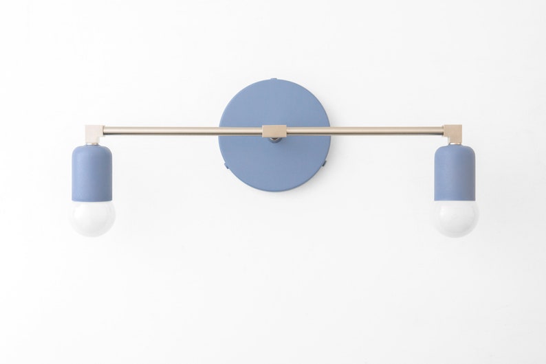 Puede incluir: Una l&aacute;mpara de pared con dos bombillas. La l&aacute;mpara tiene un brazo de metal plateado y una base redonda de color azul claro. Las bombillas est&aacute;n encerradas en cilindros de color azul claro.