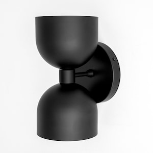 May include: A modern black wall sconce with a unique hourglass shape. The fixture features a circular base, a central connecting arm, and two rounded, bowl-shaped shades. The sconce is mounted on a white wall, creating a striking contrast.