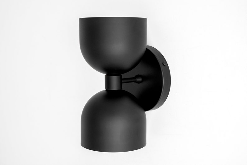 May include: A modern black wall sconce with a unique hourglass shape. The fixture features a circular base, a central connecting arm, and two rounded, bowl-shaped shades. The sconce is mounted on a white wall, creating a striking contrast.