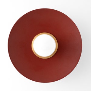 May include: A modern, circular wall sconce with a deep red finish. The sconce features a white center light surrounded by a gold-colored ring. The fixture is mounted on a white wall, creating a striking contrast.